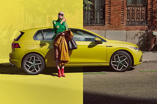Kobieta opiera się o żółtego Volkswagena na tle ceglanego budynku.